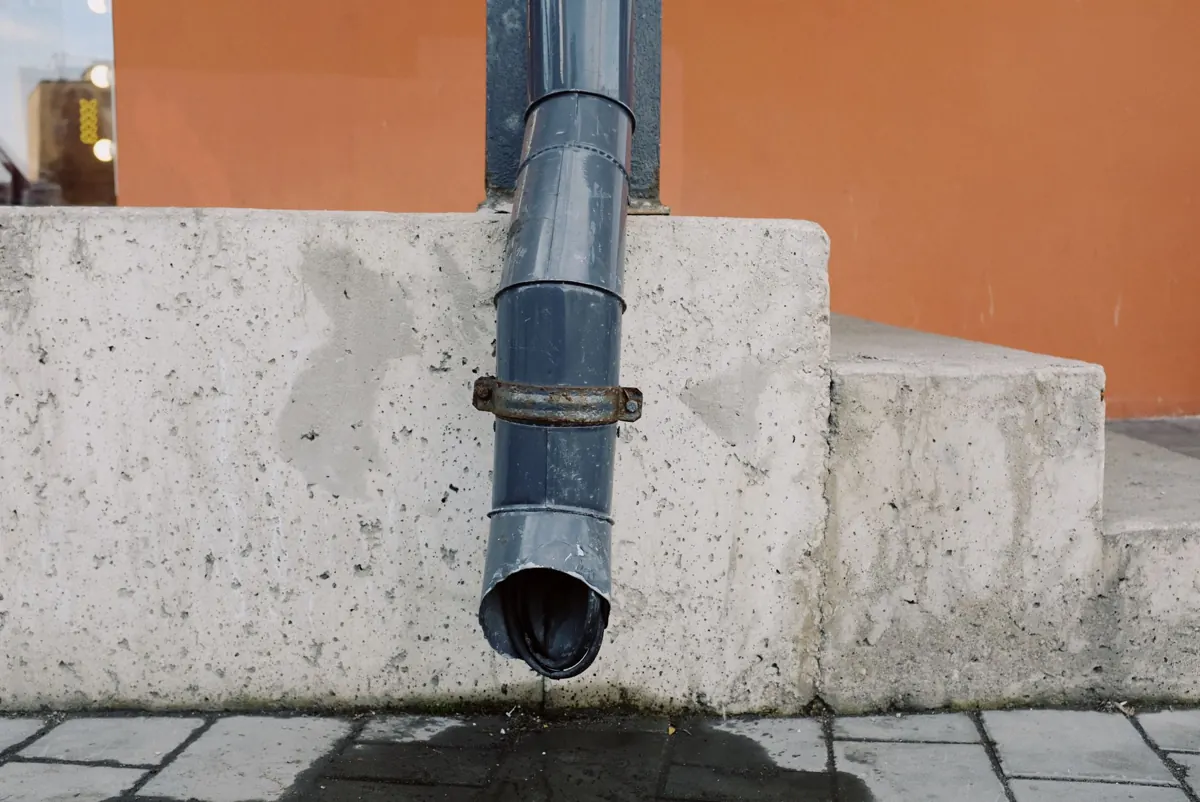 Fallrohr aus Metall an einer Hauswand, darunter eine nasse Stelle vom Regenwasser